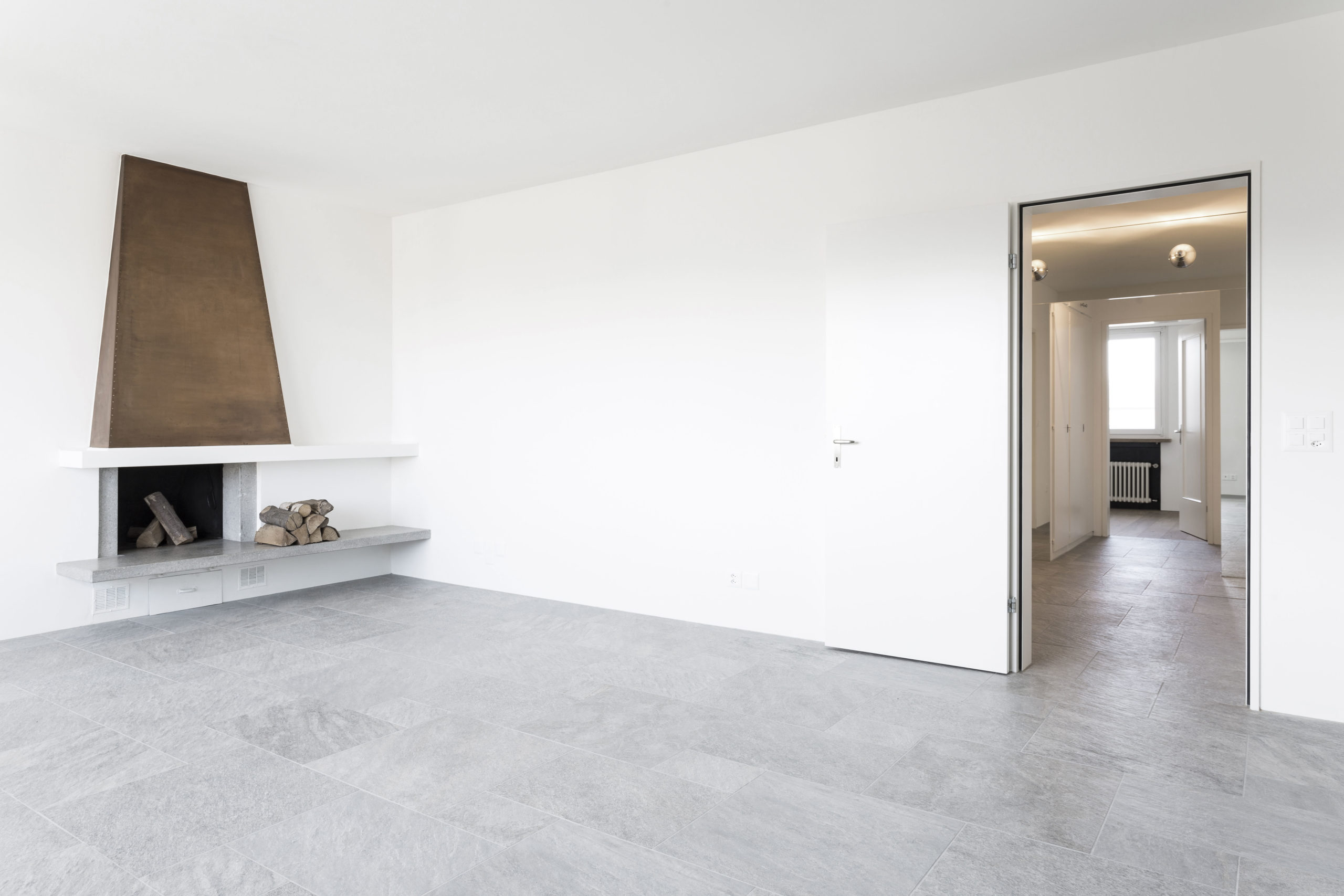 Modernes Wohnzimmer mit grauem Steinfußboden und weißer Wand, featuring eine minimalistische Kaminumrahmung mit Brennholz und eine offene Tür, die zu einem weiteren Raum führt.