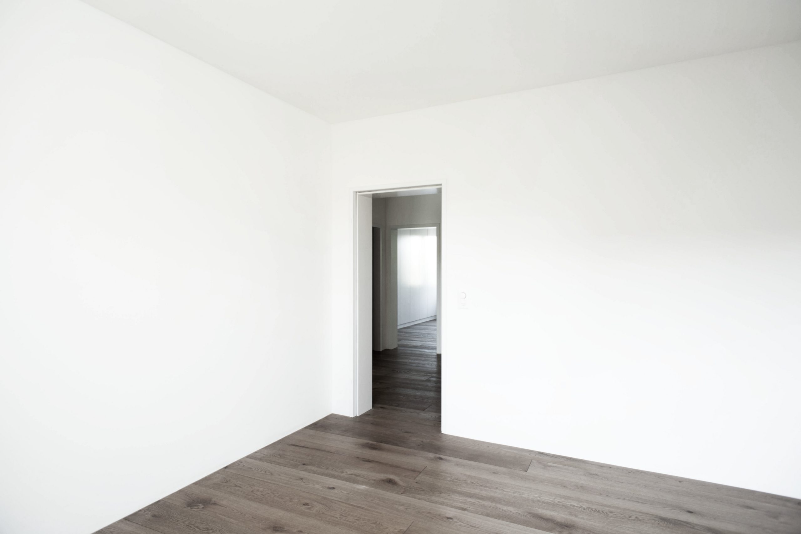 Minimalistisches leeres Zimmer mit weißen Wänden und dunklem Holzboden, offene Tür zu einem weiteren Raum.