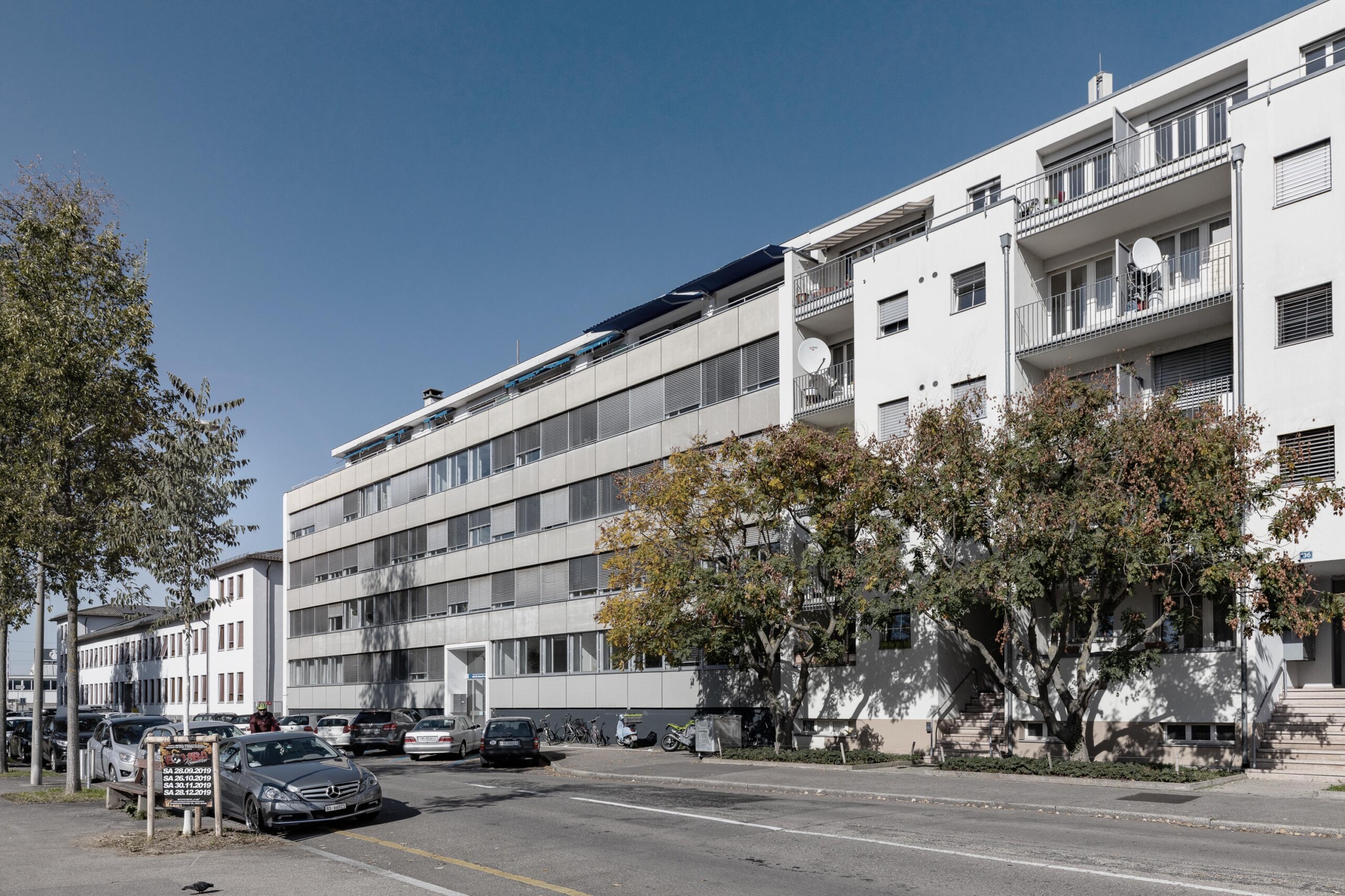 Umbau, Umnutzung und Wettbewerb von KGRUPPE Architekten Basel - Bestehendes Bürogebäude an der Hochbergerstrasse 158 mit parkenden Autos und Bäumen im Vordergrund.