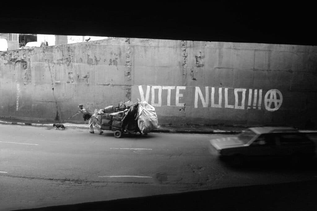 von KGRUPPE Architekten Basel - Schwarzweiss-Foto einer Strasse, auf der eine Person einen schweren Wagen und einen Hund zieht. An der Wand steht in grossen Buchstaben „VOTE NULO!!!“ und es ist ein Anarchie-Symbol zu sehen.