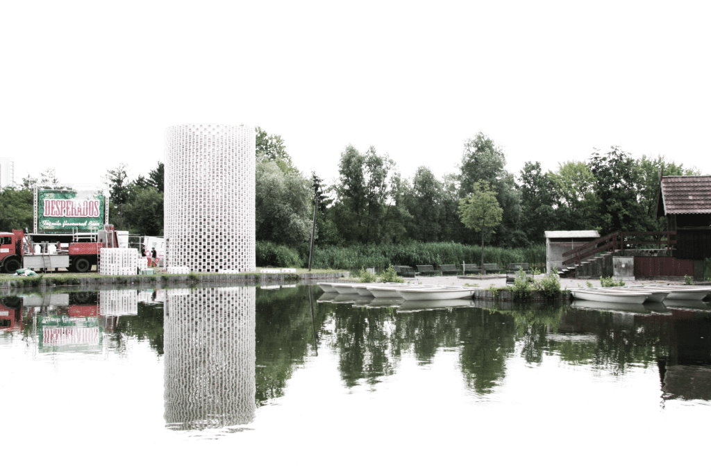 Szenografie, Umnutzung und Upcycling von KGRUPPE Architekten Basel - Zylinderförmige Struktur und Boote an einem reflektierenden See vor einem Waldrand.