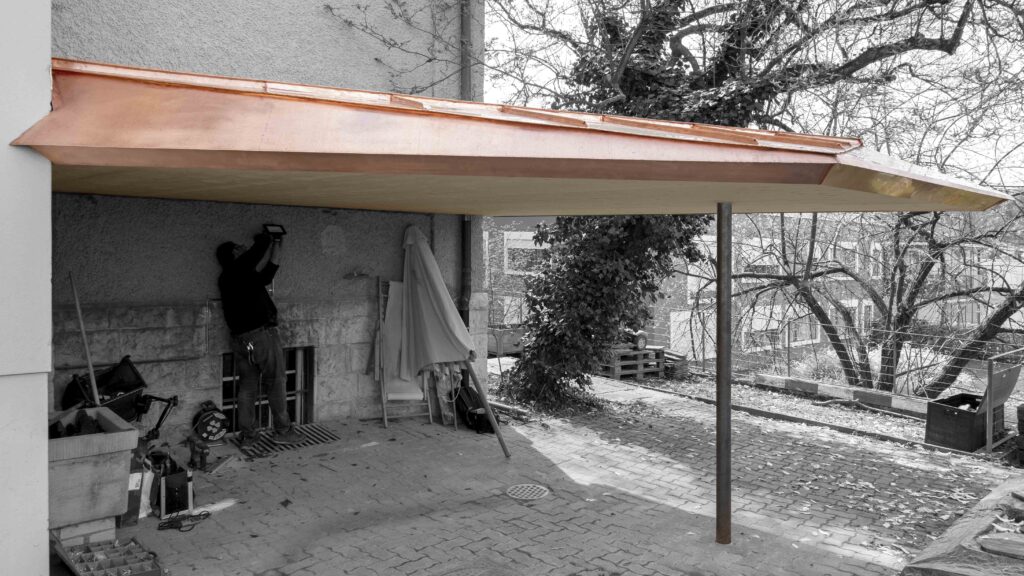 Neubau und Szenografie von KGRUPPE Architekten Basel - Ein Handwerker installiert ein Licht unter einem Carport mit Kupferdach, das an einem Haus befestigt ist. Im Hintergrund sind Bäume und eine gepflasterte Auffahrt zu sehen.