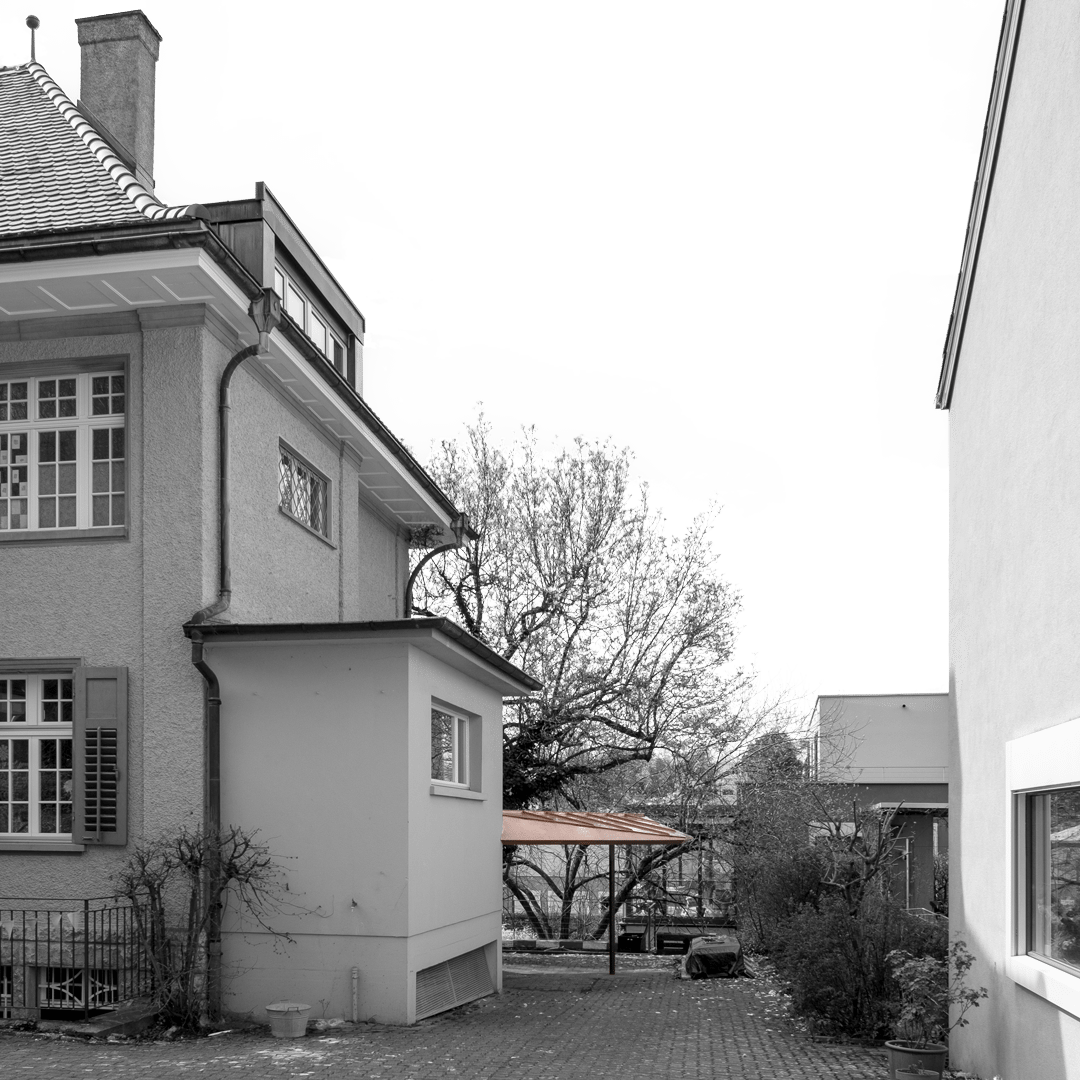 Schwarz-Weiss-Fotografie eines Wohnhauses mit mehreren Fenstern und einem kleinen Anbau. Ein schmaler Weg trennt das Haus von einem weiteren Gebäude auf der rechten Seite. Im Hintergrund sind kahle Bäume und ein Carport zu sehen.