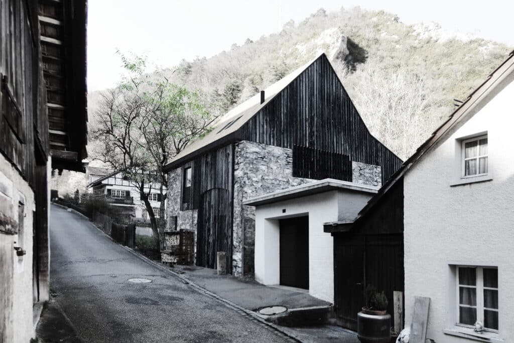 Neubau, Umbau und Umnutzung von KGRUPPE Architekten Basel - Enge Gasse mit alten Steinhäusern und Scheunen, umgeben von hügeliger Landschaft.