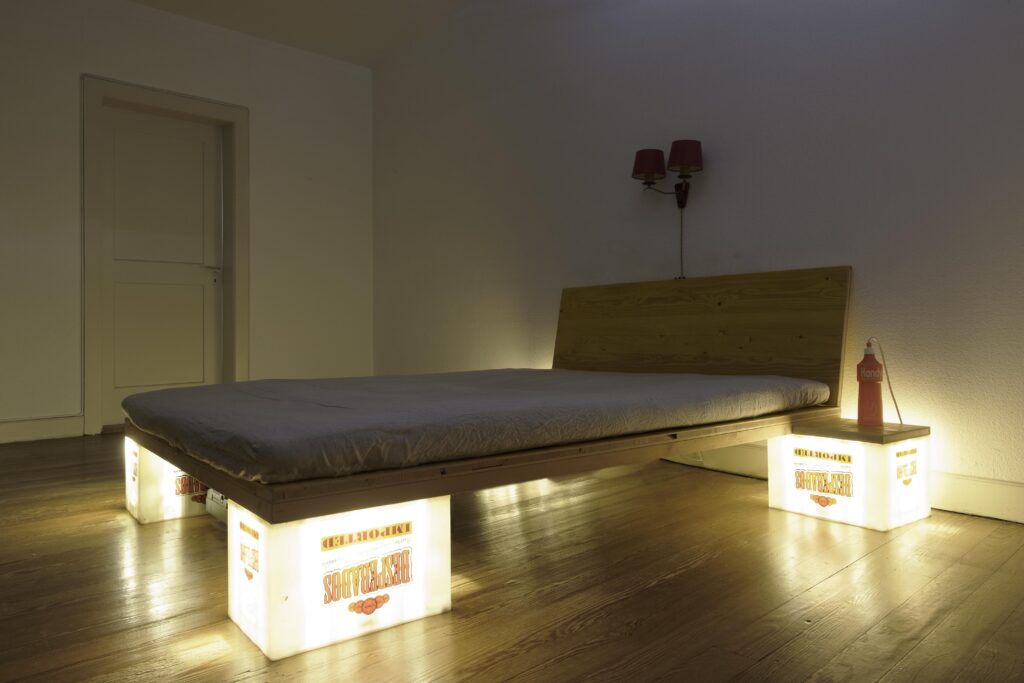 Leuchte, Möbel und Upcycling von KGRUPPE Architekten Basel - Minimalistisch eingerichtetes Schlafzimmer mit einem Bett, das auf leuchtenden Getränkekisten steht.