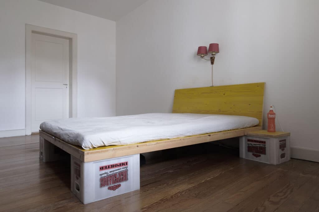 Leuchte, Möbel und Upcycling von KGRUPPE Architekten Basel - Ein einfaches Bett mit einem hölzernen Kopfteil, das auf Kunststoffkisten steht, in einem minimalistisch eingerichteten weissen Raum mit Holzboden.