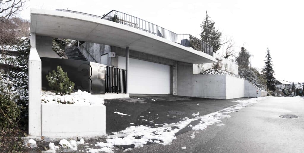 Neubau und Umbau von KGRUPPE Architekten Basel - Garageneinfahrt eines modernen Hauses mit Betonwänden, schwarzem Tor und der Hausnummer 3 im verschneiten Umfeld.