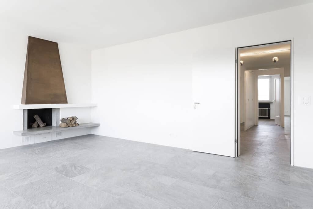 von KGRUPPE Architekten Basel - Renovation eines Penthouses durch das Architekturbüro KGRUPPE Architekten Basel. Ein klassisch modernes, leeres Wohnzimmer mit grauem Fliesenboden, weiss gestrichenen Wänden und einem abstrakten Kamin.