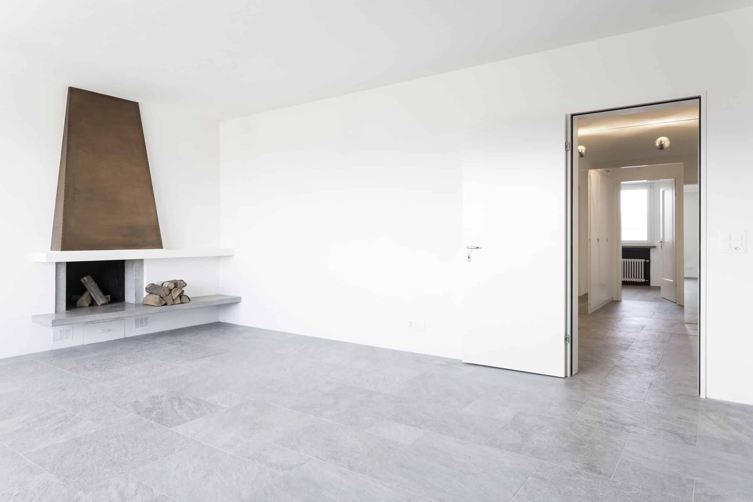 Renovation eines Penthouses durch das Architekturbüro KGRUPPE Architekten Basel. Ein klassisch modernes, leeres Wohnzimmer mit grauem Fliesenboden, weiss gestrichenen Wänden und einem abstrakten Kamin.