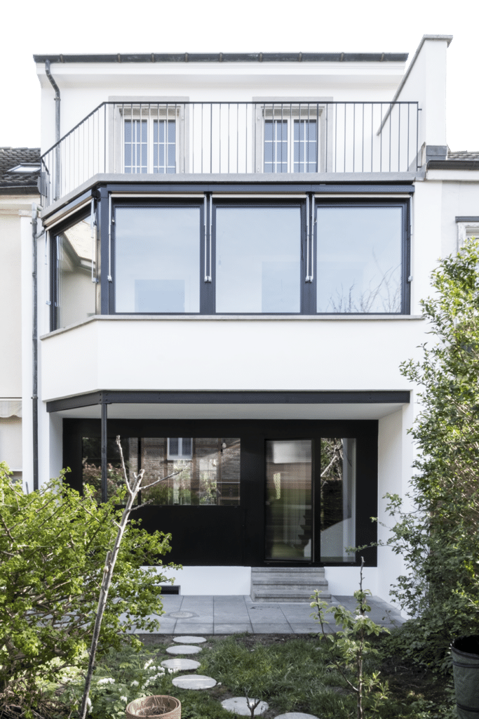 von KGRUPPE Architekten Basel - Umbau eines Einfamilienhauses durch das Architekturbüro KGRUPPE Architekten Basel. Modernes zweistöckiges Haus mit Glasfassade und Balkon, umgeben von einem Garten mit gepflastertem Weg.