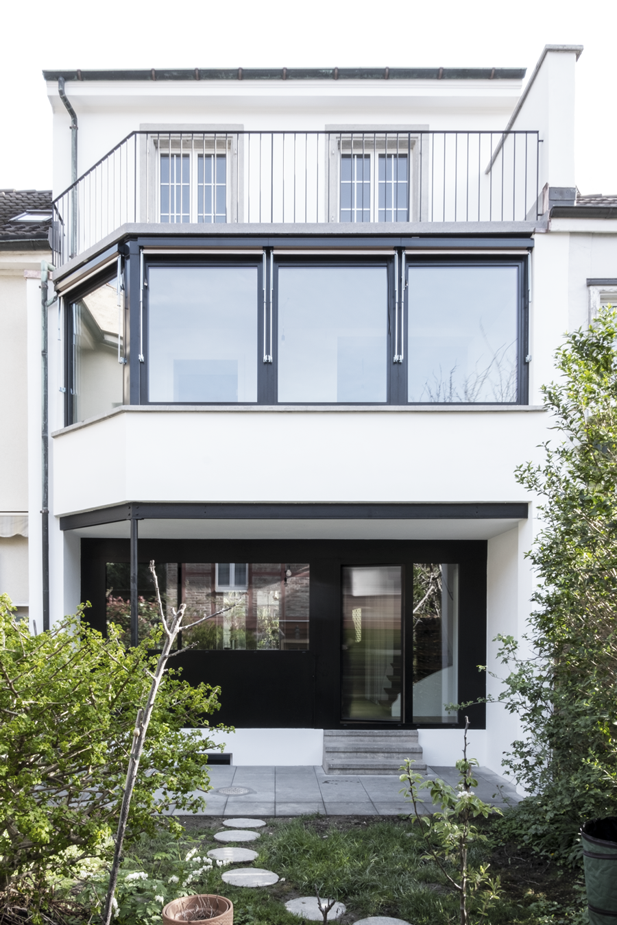 Umbau eines Einfamilienhauses durch das Architekturbüro KGRUPPE Architekten Basel. Modernes zweistöckiges Haus mit Glasfassade und Balkon, umgeben von einem Garten mit gepflastertem Weg.