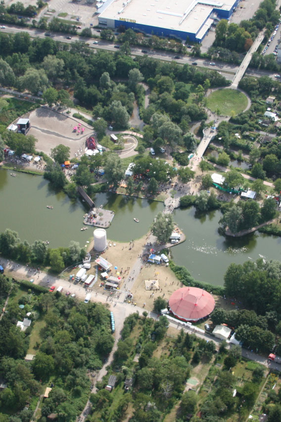 Szenografie, Umnutzung und Upcycling von KGRUPPE Architekten Basel - Luftaufnahme eines Parks mit Teichen, Bäumen, Gehwegen und zahlreichen Zelten und Ständen.