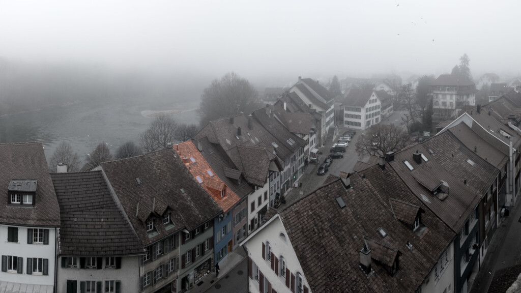 von KGRUPPE Architekten Basel - Nebliger Blick auf ein Flussuferstädtchen mit dicht aneinanderstehenden Satteldächern; ein Dach mit orangefarbenen Ziegeln sticht hervor.