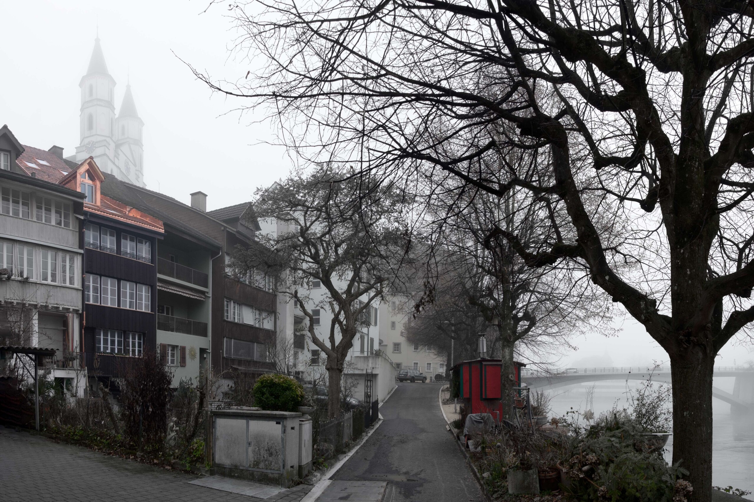 von KGRUPPE Architekten Basel - Nebliger Flussweg mit kahlen Bäumen, alten Wohnhäusern, rotem Kiosk am Ufer und verschwommenen Kirchtürmen.