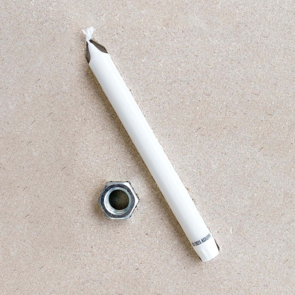  von KGRUPPE Architekten Basel - Weisse Kerze mit abgebrannter, verkohlter Spitze und sichtbarem Docht liegt diagonal auf einem hellen, strukturierten Untergrund neben einer silbernen Sechskantmutter.