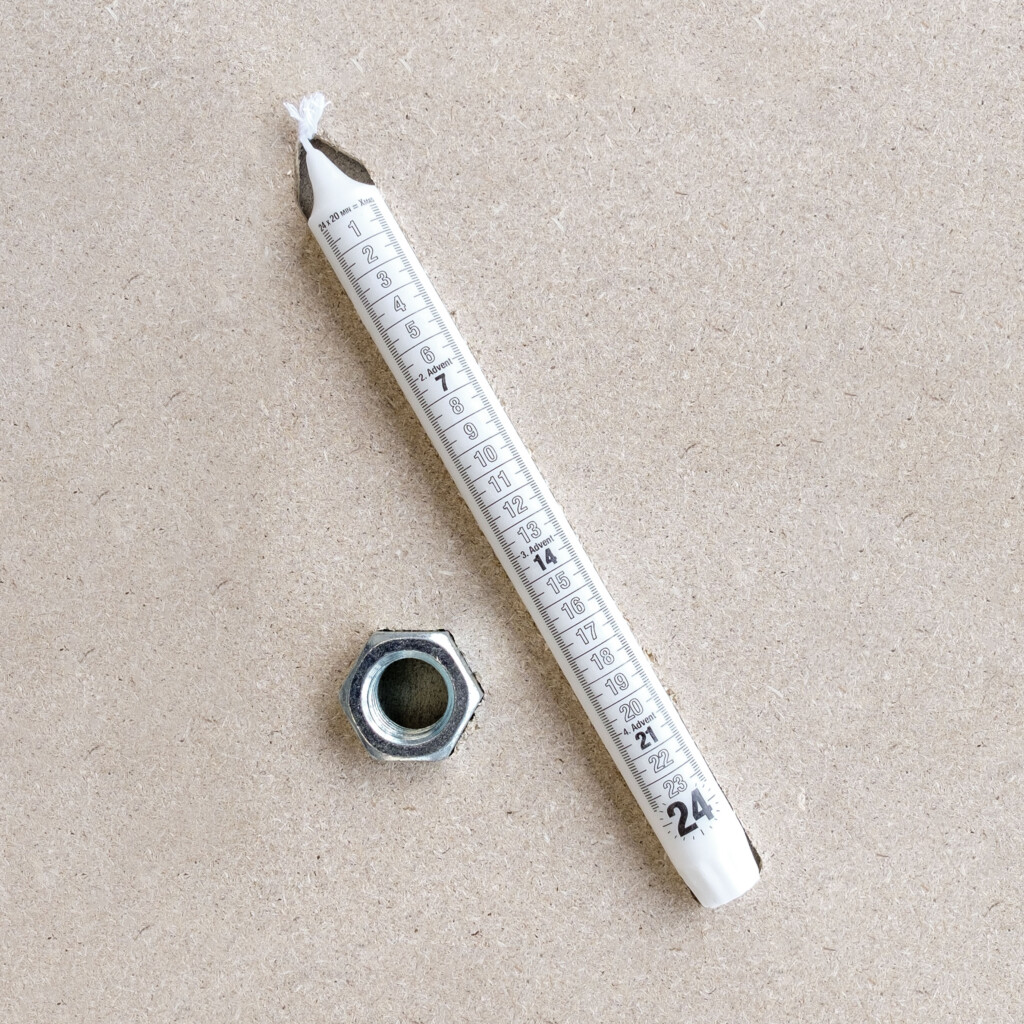  von KGRUPPE Architekten Basel - Weiße Adventskerze mit aufgedruckten Zahlen 1–24 und sichtbarem Docht, schräg auf einer hellen Holzpressplatte liegend, daneben eine sechseckige Metallmutter.