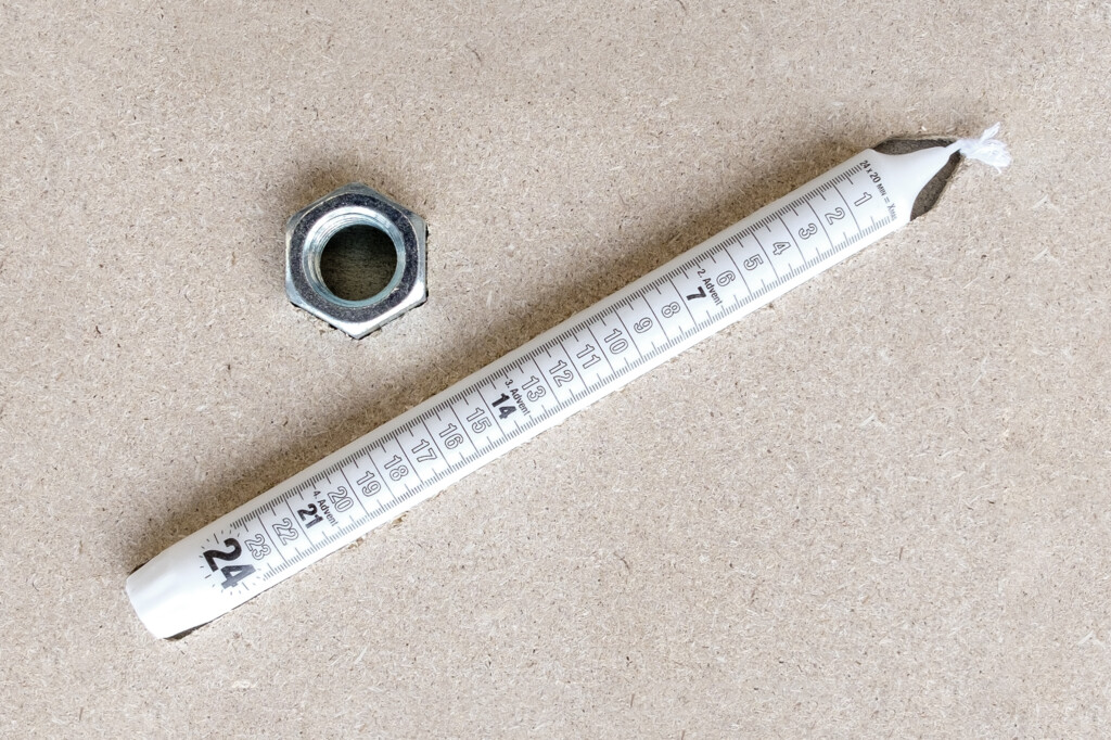 von KGRUPPE Architekten Basel - Weiße Adventskerze mit aufgedruckten Zahlen 1–24 und sichtbarem Docht, schräg auf einer hellen Holzpressplatte liegend, daneben eine sechseckige Metallmutter.