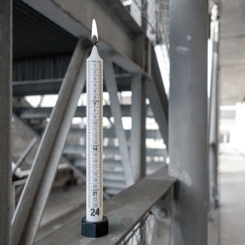  von KGRUPPE Architekten Basel - Brennende weisse Adventskerze mit aufgedruckten Zahlen 1–24 in einem schwarzen sechseckigen Halter, auf einem Geländer vor einer verschwommenen, grauen Industrietreppe positioniert.